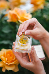 Käsi hoiab väikest parfüümipudelit, taustal kollased lilled